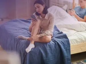Homem deitado na cama entretido ao celular enquanto a mulher está sentada aos pés da cama também entretida ao celular!