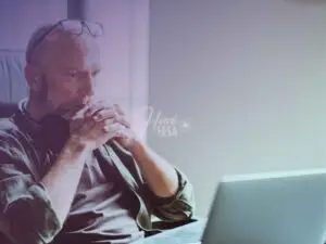 Homem de cabelos grisalhos sentado em frente ao notebook com as mãos cruzadas junto ao queixo em gesto de reflexão profunda enfatizando a crise dos 50 anos nos homens