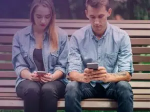 Homem e mulher sentados em um banco de praça com as pernas encostadas, dando a entender que se trata de um casal, mas ambos estão entretidos, cada um em seu celular sem dar a devida tenção um ao outro.