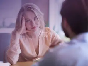 Mulher sentada à mesa com a mão direita no rosto, olhando para o lado com expressão de entediada enquanto homem fala com ela no outro lado da mesa, provavelmente uma conversa entre casal onde a mulher está desinteressada pelo marido!