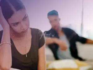 Homem sentado no sofá ao fundo da imagem olhando para a mulher em ar de tristeza devido ao fato do marido não querer sair com ela, sentada na ponta do sofá em destaque na imagem!