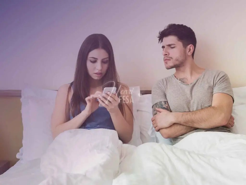 Mulher mexendo no celular, sentada e encostada na cabeceira da cama, enquanto homem, também sentado e encostado na cabeceira da cama ao lado da mulher, se estica para ver o que ela está fazendo!