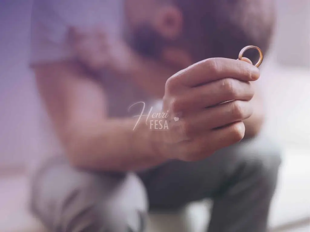 Homem sentado no sofá com a cabeça abaixada e com a mão esquerda erguida segurando a aliança após a separação!