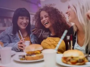 Três mulheres sentadas em uma mesa de lanchonete, comendo hambúrguer, tomando refrigerante, e todas sorrindo e rindo com intensidade, mostrando que estão felizes!