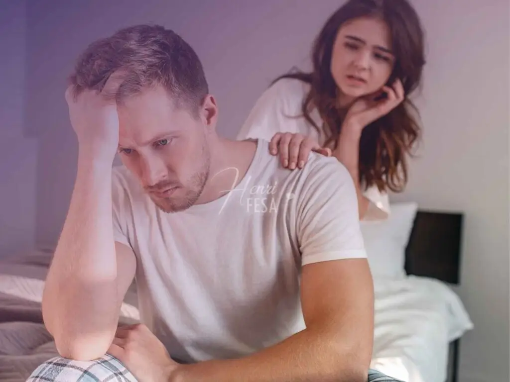 Homem sentado nos pés da cama com o cotovelo no joelho e a mão na cabeça em tom de reflexão e desânimo, enquanto mulher tenta consolá-lo colocando a mão em seu ombro!