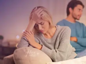 Mulher sentada no sofá, com as mãos na cabeça e os olhos fechados, expressando descontentamento profundo, enquanto homem está sentado na outra extremidade do mesmo sofá, com os braços cruzados em expressão de reflexão.