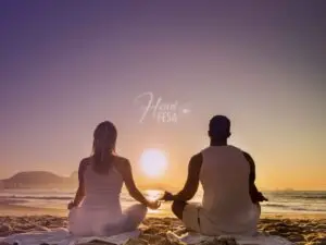 Casal, homem e mulher, sentados juntos em posição de meditação, ambos de frente para o mar em um belo por do sol!
