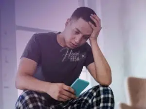 Homem triste, sentado, com uma mão da cabeça e outra segurando um celular.