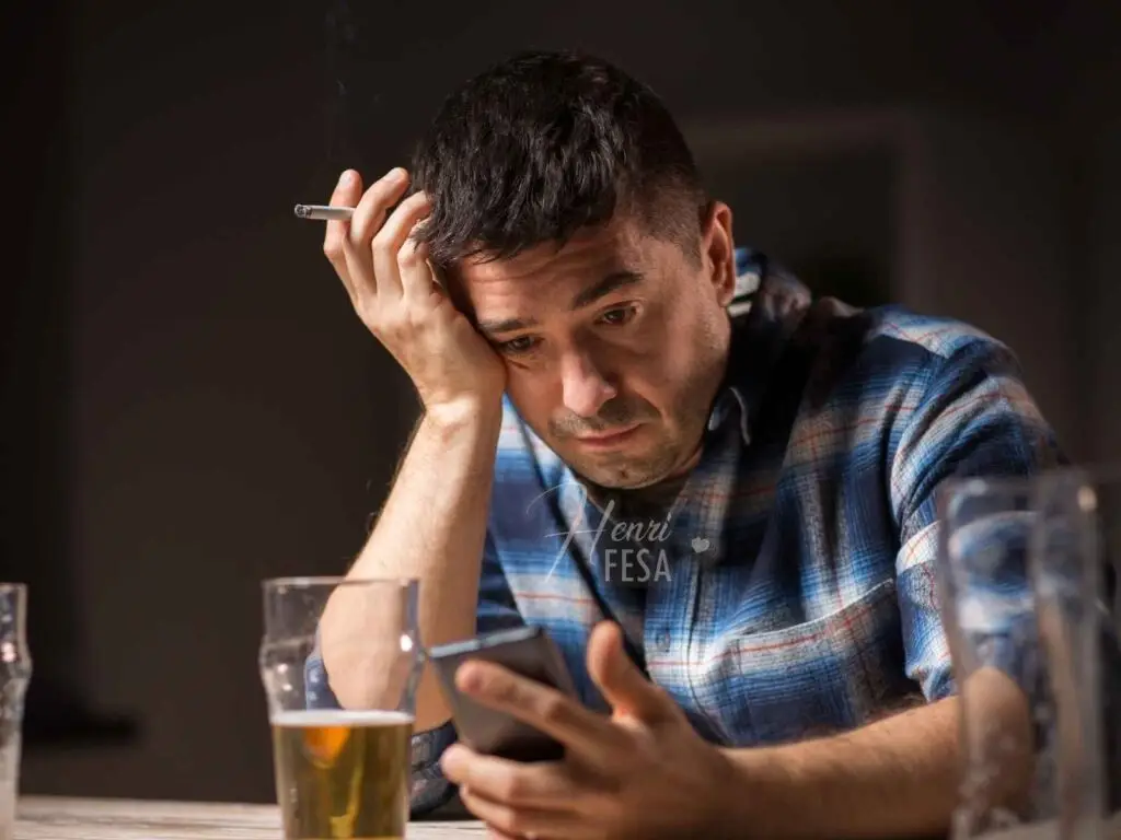 Homem alcoolizado, sentado à mesa em frente a um copo de cerveja, mexendo no celular com um semblante triste!