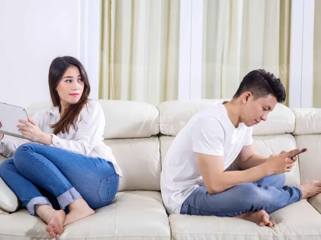 Mulher olhando desconfiada para o marido que está de costas para ele todo sorridente mexendo no celular!