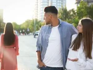 Homem virando o pescoço e olhando uma mulher passando enquanto abraçado a sua parceira. A parceira dele está olhando indignadamente para ele enquanto ele olha para outra!