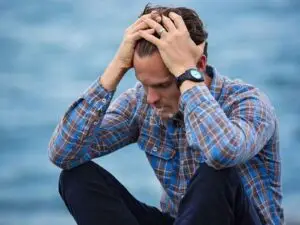 Homem sentado à beira do lago, com as mãos na cabeça de pensativamente com semblante de frustração evidente!