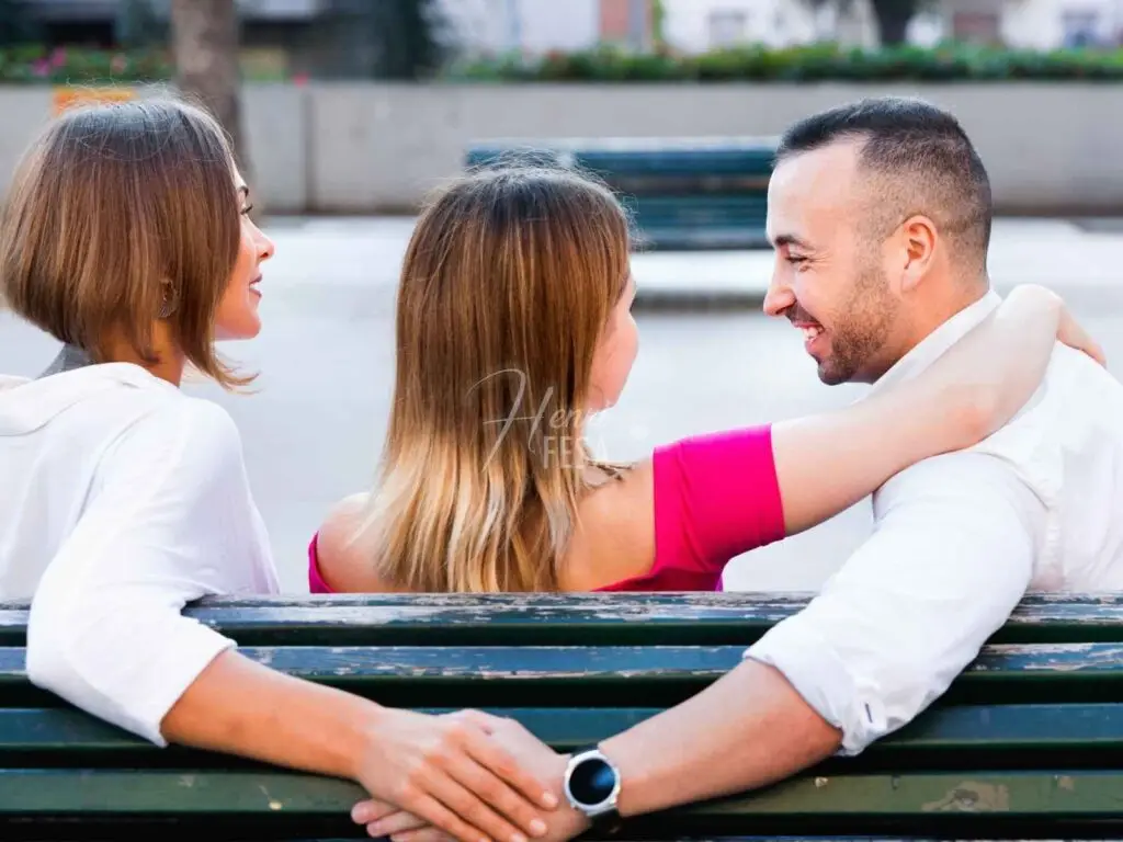 Casal sentado no banco da praça abraçados, amiga do casal sentada logo ao lado, enquanto o marido abraça a esposa e segura a mão da amiga simultaneamente, sem que a esposa perceba em um ato de atração por outra pessoa fora do casamento!