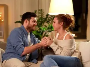 Casal sentado ao sofá, de mão dadas e ambos com um belo sorriso no rosto, ambos se olhando apaixonados, sendo essa uma das coisas para um relacionamento dar certo!