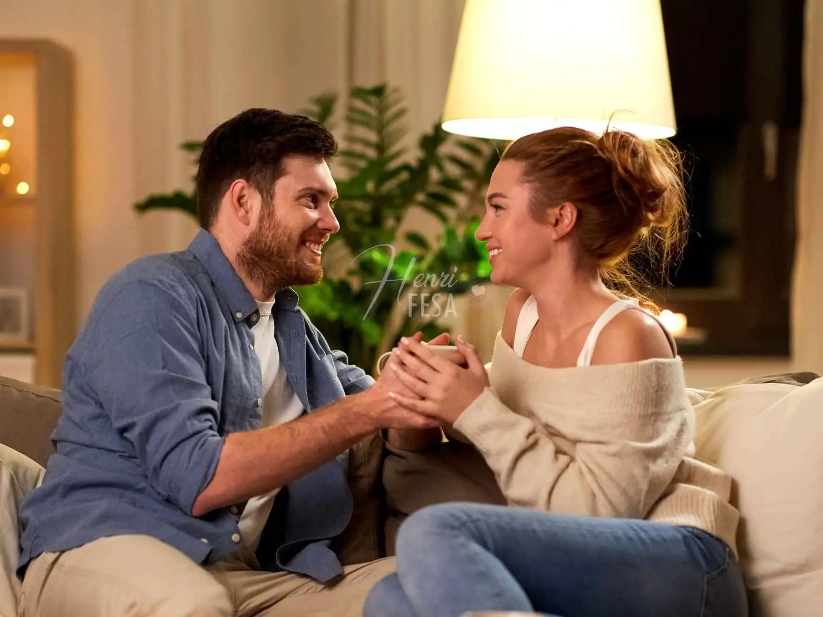 Casal sentado ao sofá, de mão dadas e ambos com um belo sorriso no rosto, ambos se olhando apaixonados, sendo essa uma das coisas para um relacionamento dar certo!