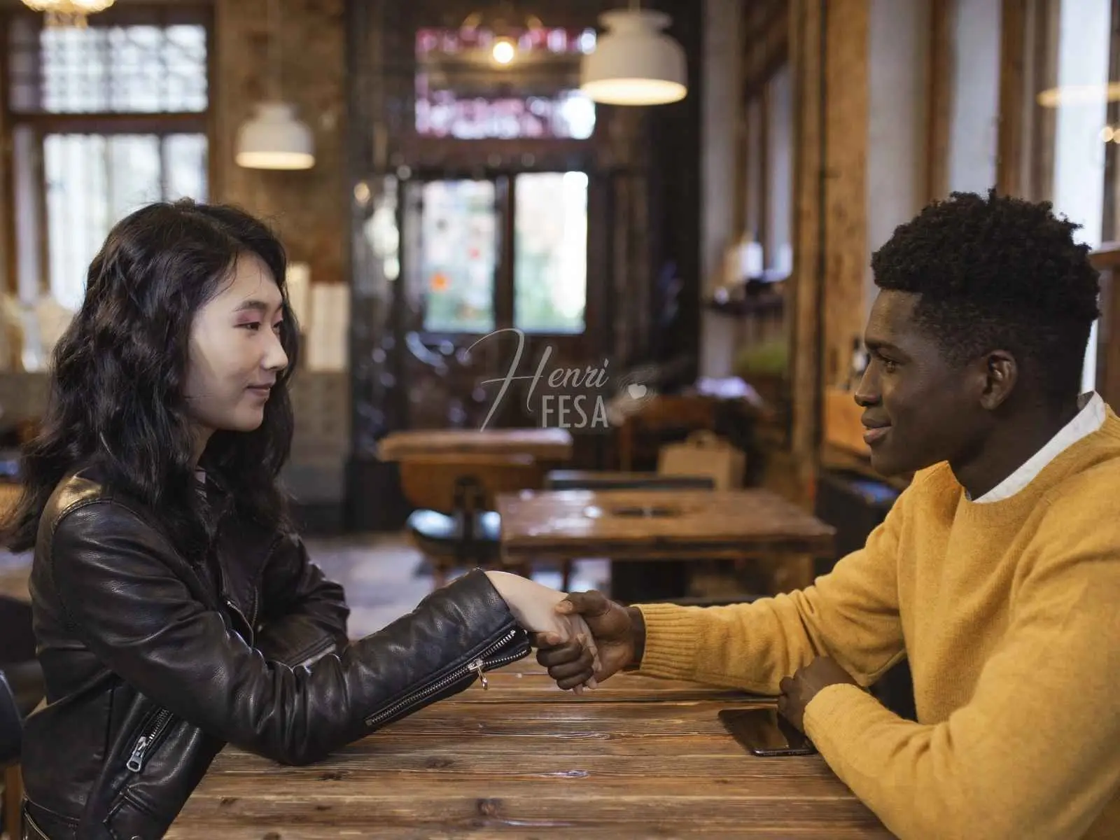 Homem e mulher sentados em uma mesa de restaurante/bar, ambos um de frente para o outro, ambos sorrindo e segurando as mãos, um ato de como atiçar uma mulher com toque, gestos e sorrisos!