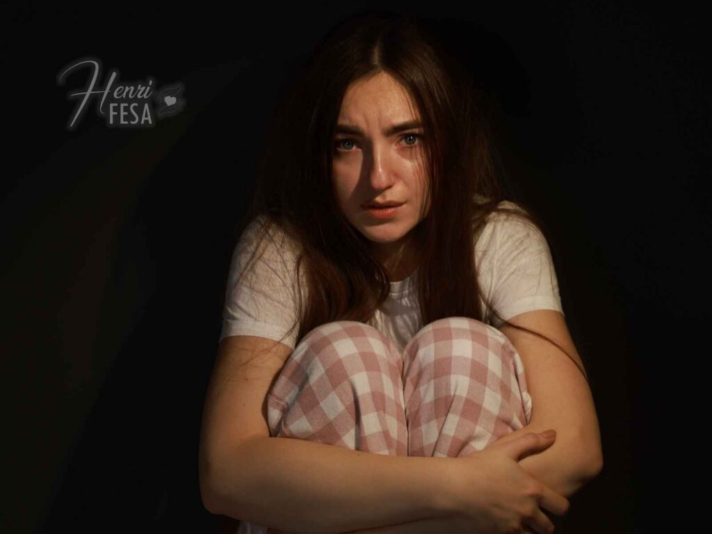 Como lidar com o medo de ter filhos? Mulher agachada com as mãos nos joelhos, com semblante de medo e fundo totalmente escuro, indicando um medo de ter filhos em grau de fobia extrema!