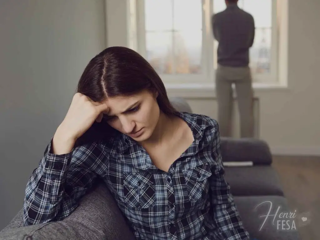 Quando o casal não tem os mesmos objetivos? Mulher pensativa a beira do sofá, homem logo atrás olhando pela janela, também pensativo quando o casal não tem os mesmos objetivos e diverge!