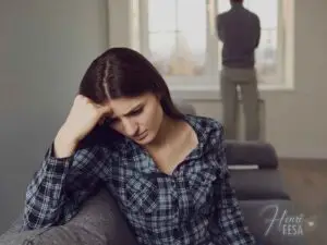 Mulher pensativa a beira do sofá, homem logo atrás olhando pela janela, também pensativo quando o casal não tem os mesmos objetivos e diverge!