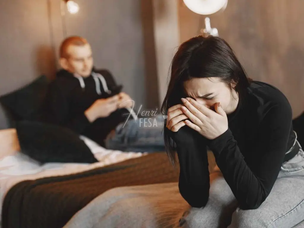 Sinais de que ele não te quer mais: saiba quais! Homem sobre a cama encostado na cabeceira, sorrindo e mexendo no celular, mulher chorando com as mãos no rosto a beira da cama, percebendo de forma evidente os sinais de que ele não te quer mais!